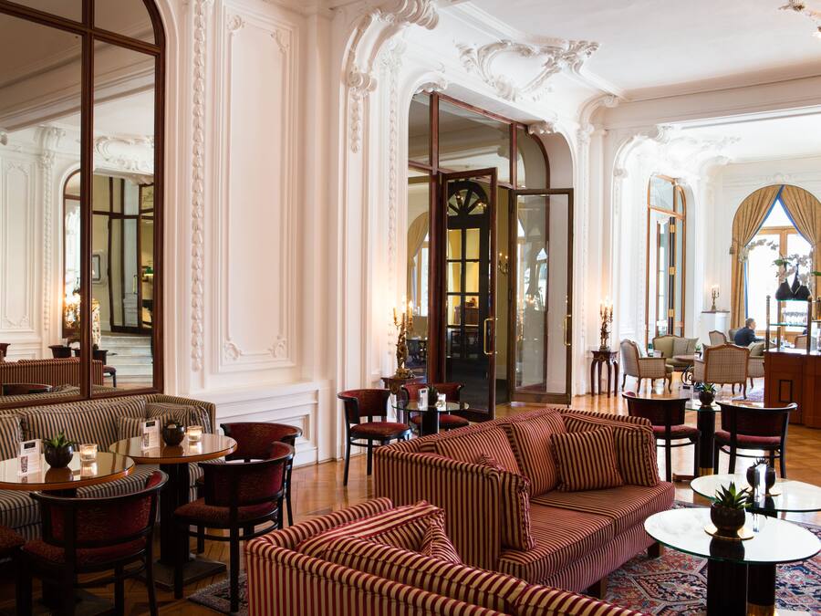 Un lobby de hotel lujoso con paredes blancas ornamentadas, una escalera monumental y un área de estar con sofás y sillones a rayas.