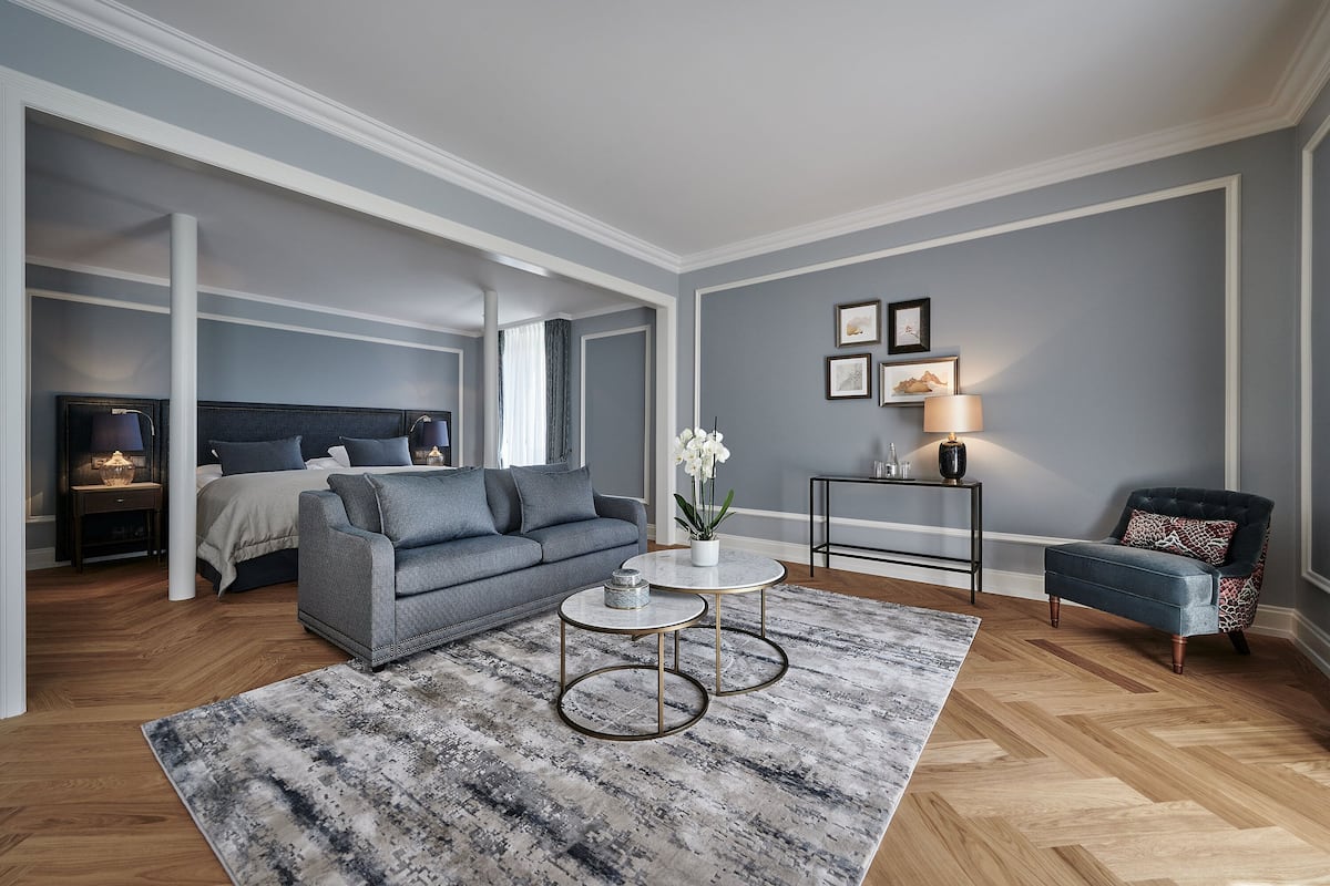 Un dormitorio moderno con un sofá gris, una cama con cabecera blanca, una mesita redonda con una lámpara y un sillón azul.