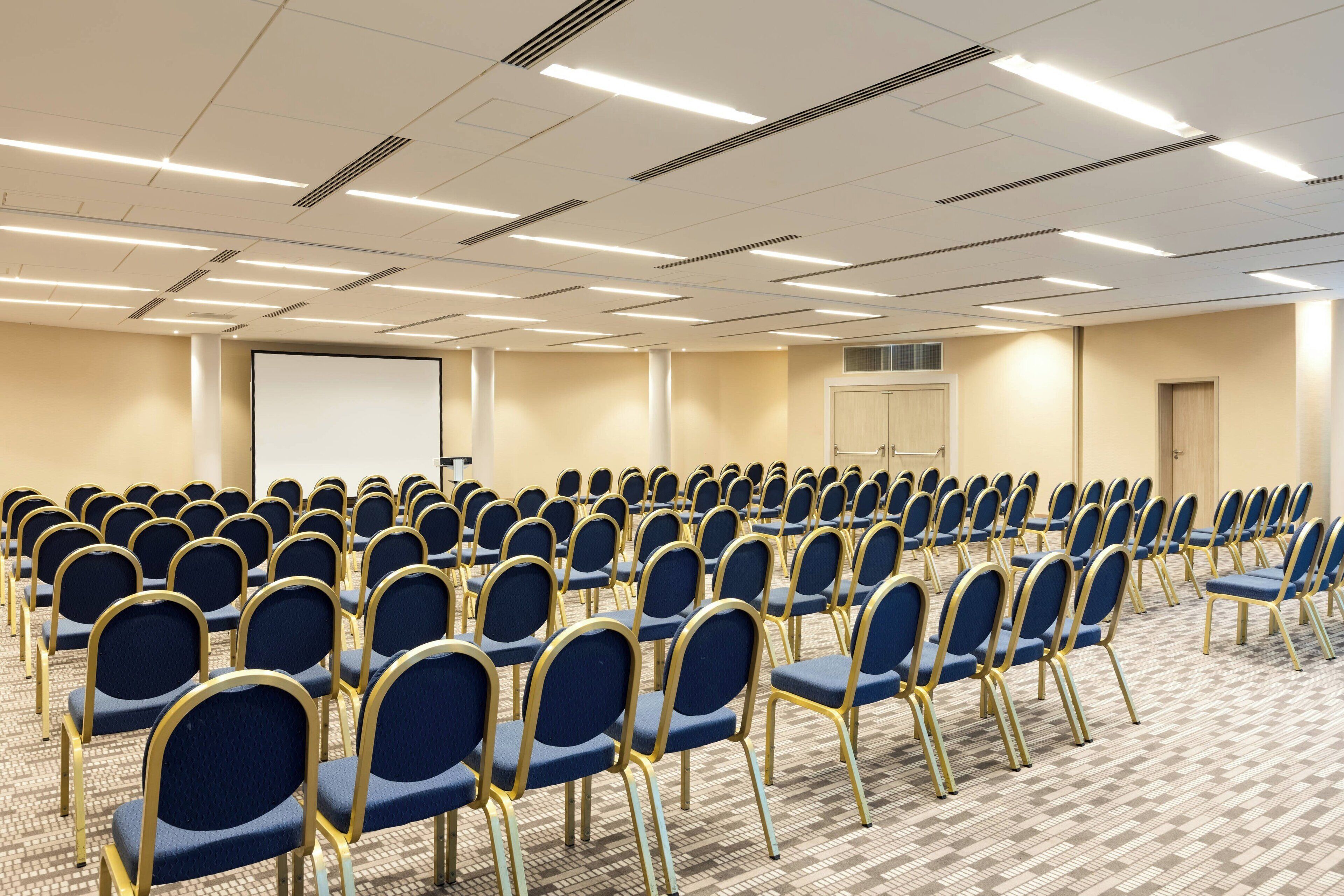 A conference room with rows of blue chairs and a projector screen.