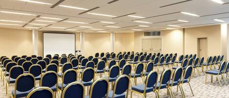 Un salón de conferencias con filas de sillas azules y una pantalla para proyector.