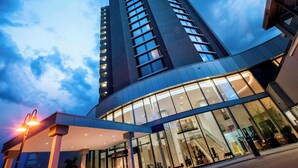 A modern hotel with a curved glass facade, a prominent entrance with a staircase, and a tall building in the background. - Delta Hotels by Marriott Frankfurt Offenbach (Offenbach am Main)