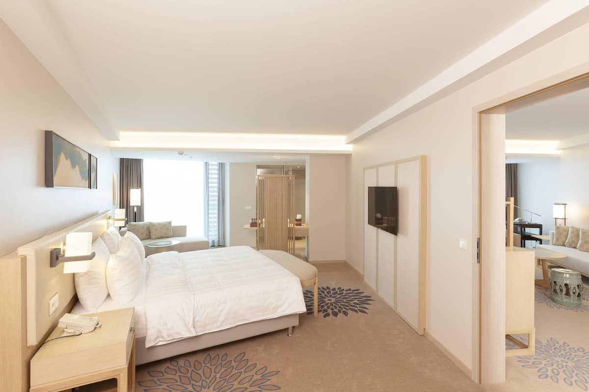 A modern hotel room with a large bed, bedside tables, a flat-screen TV, and a view of the adjacent room.