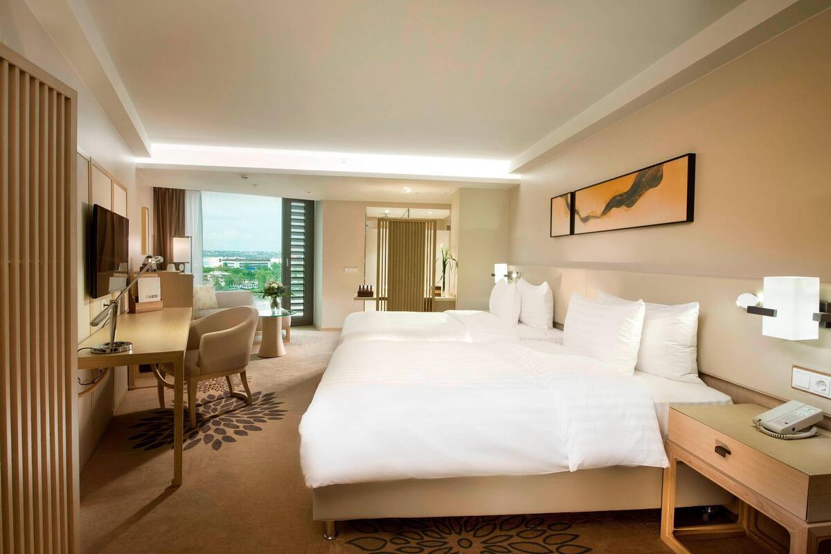 A hotel room with a large bed, a desk, a chair, a telephone, and a window with a view of the city.