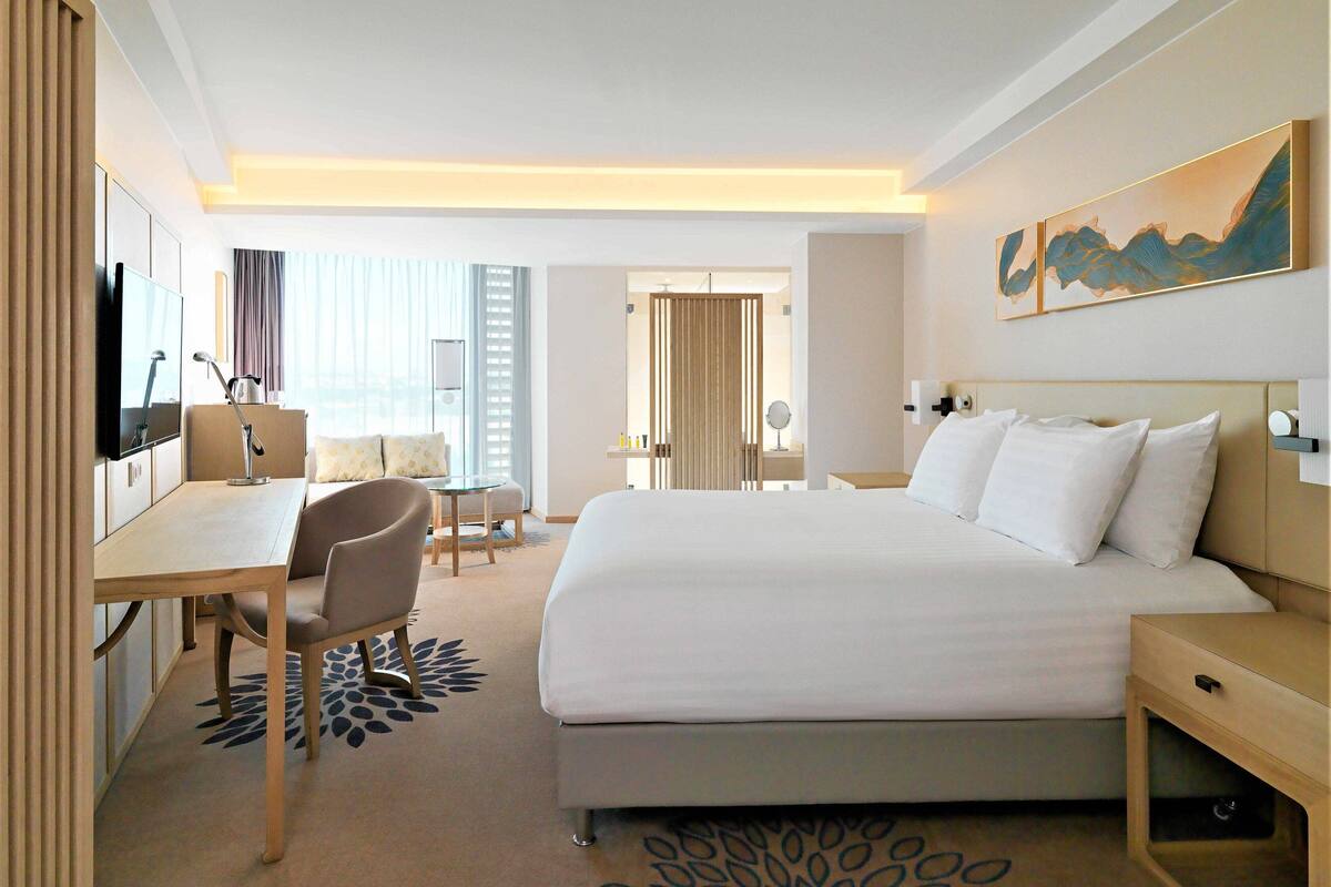 A hotel room with a bed, a desk, a chair, a TV, and a painting on the wall.