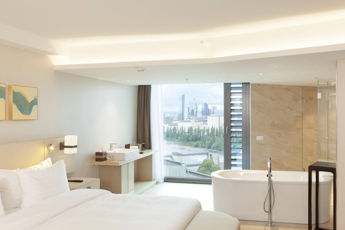 Ein modernes Hotelzimmer mit einem großen Bett, einem Badezimmer mit Badewanne, einem Schreibtisch und Blick auf die Stadt.