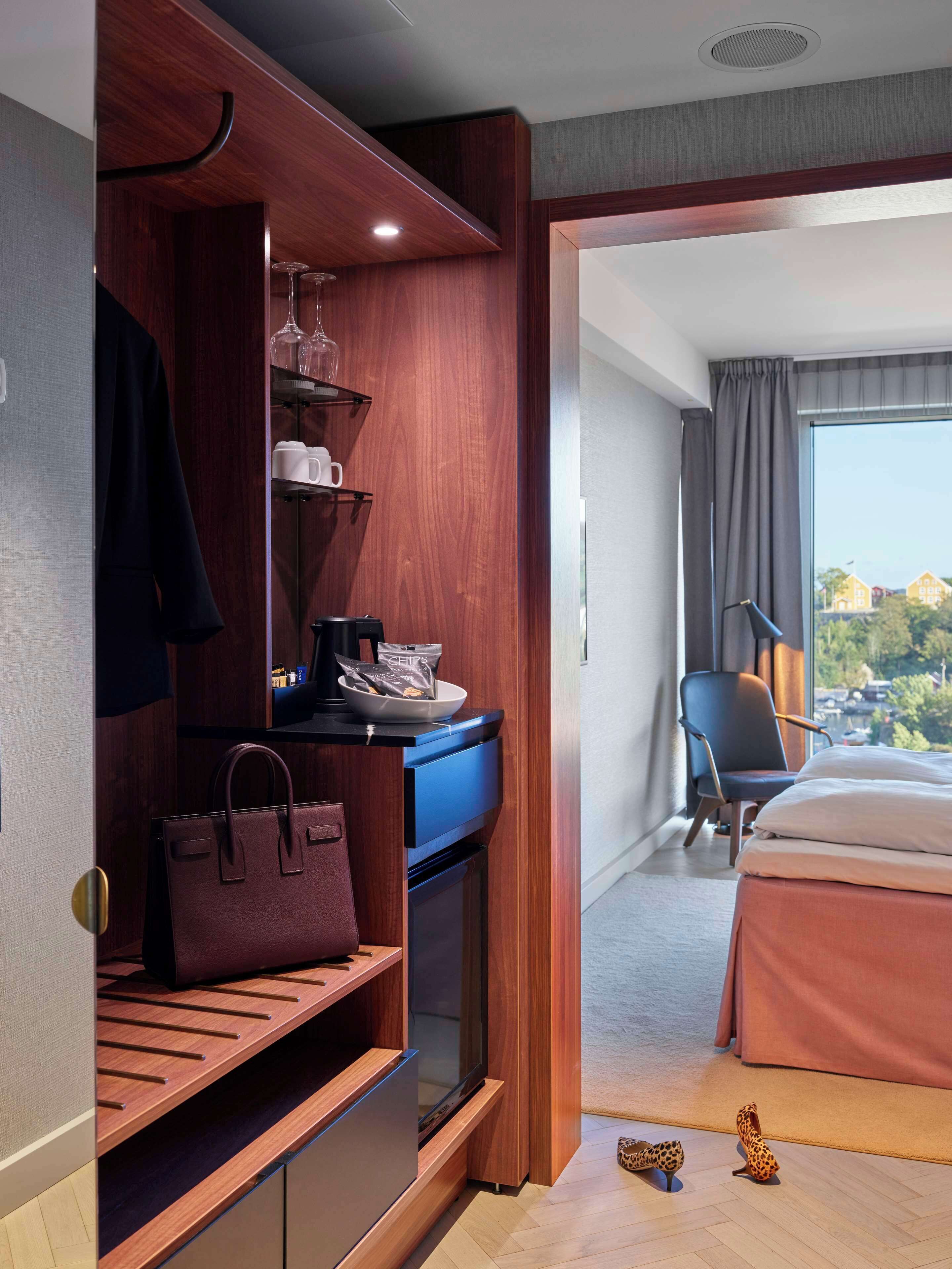 A hotel room with a wooden wardrobe, a bed, a desk with a chair, and a view of the city through the window.