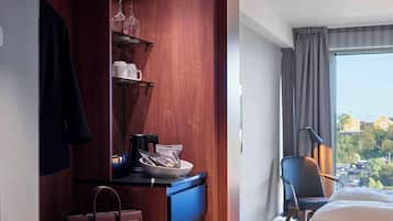 A hotel room with a wooden wardrobe, a bed, a desk with a chair, and a view of the city through the window.