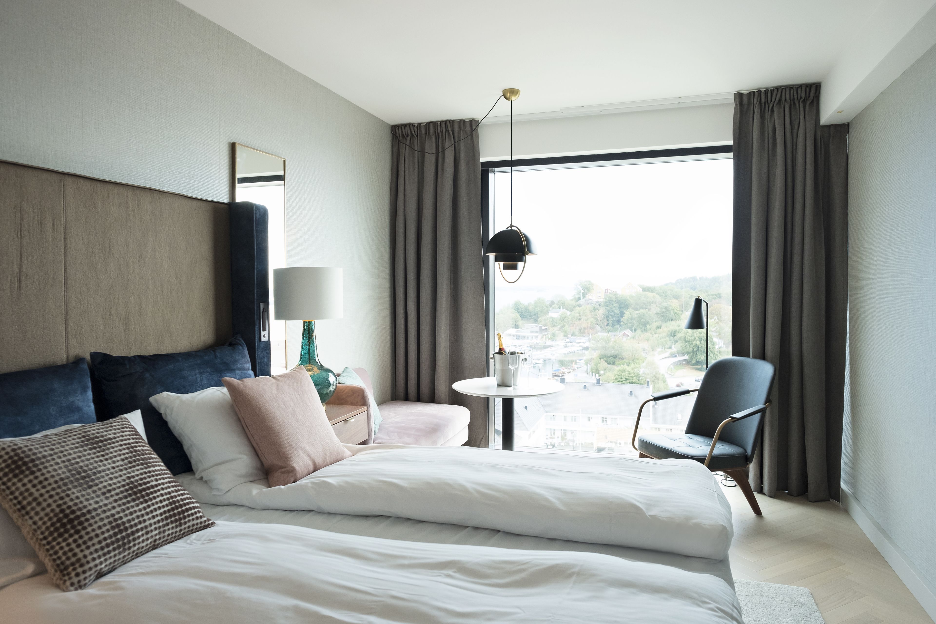 A modern hotel room with a large bed, a chair, a small table, and a view of the city through large windows.