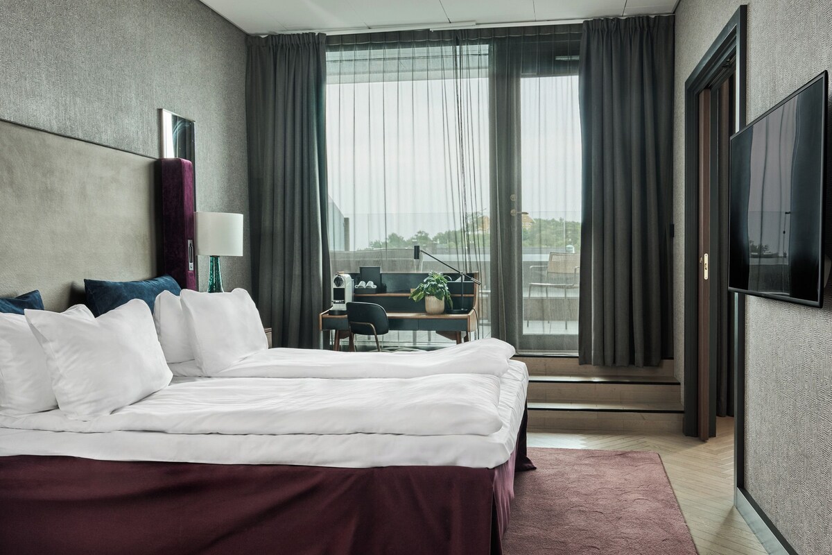 A modern hotel room with a large bed, a desk with a chair, a TV, and a view of the city.