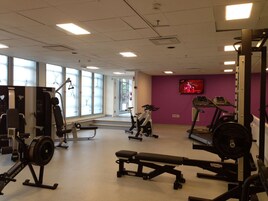 Un gimnasio con diversos equipos de ejercicio, ventanas amplias y un televisor en la pared.