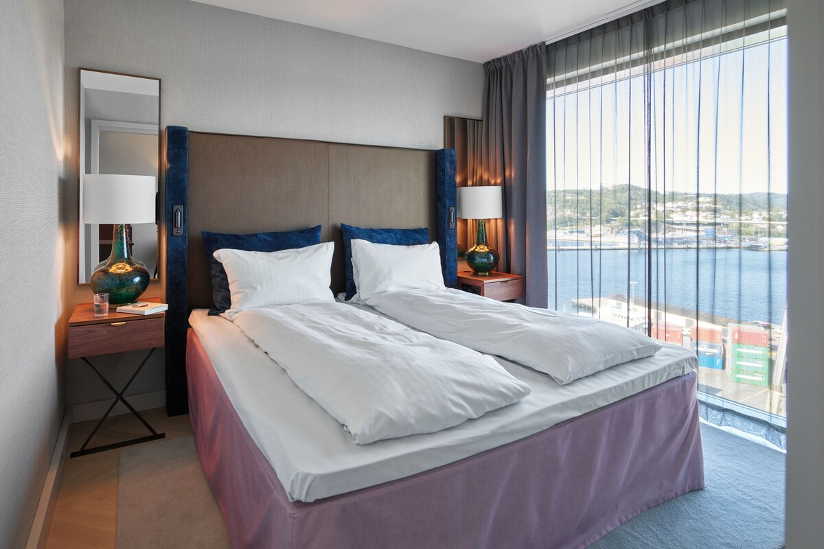 A hotel room with a large bed, two bedside tables with lamps, a nightstand, a mirror, and a view of the sea.