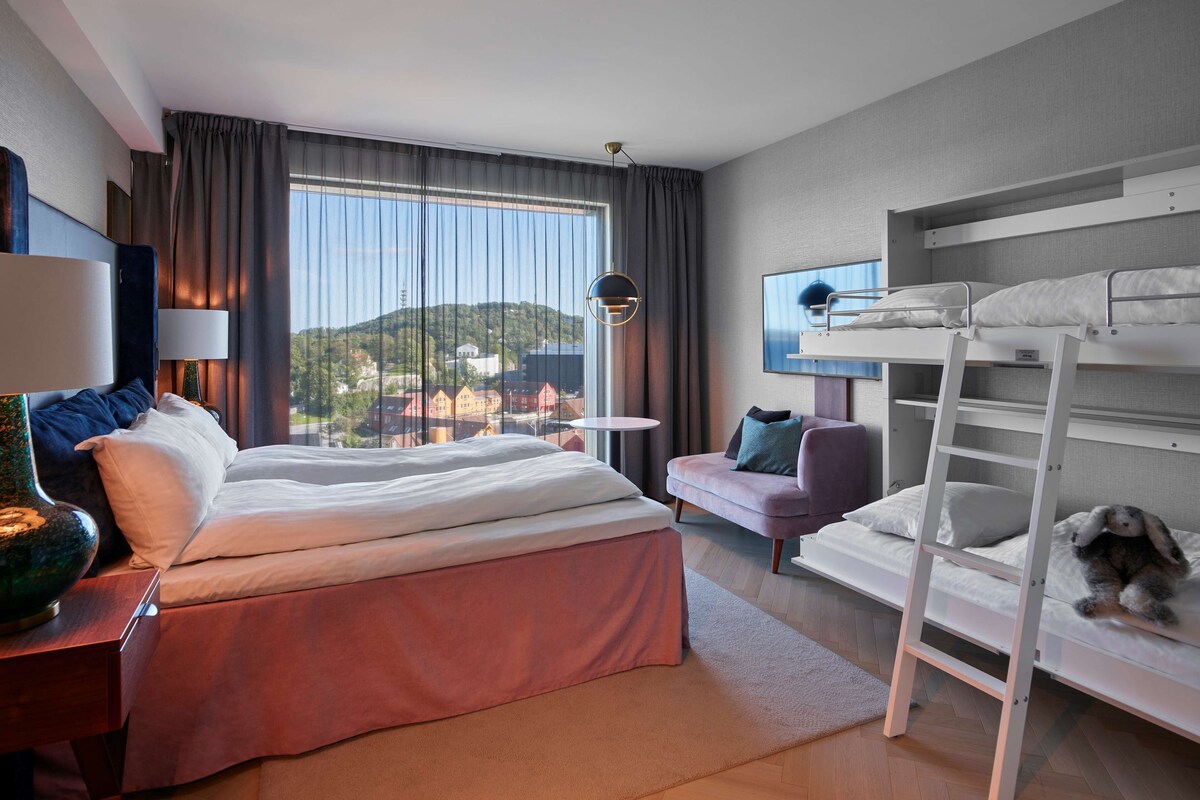 A hotel room with a large bed, a sofa, a bunk bed, and a view of a cityscape.