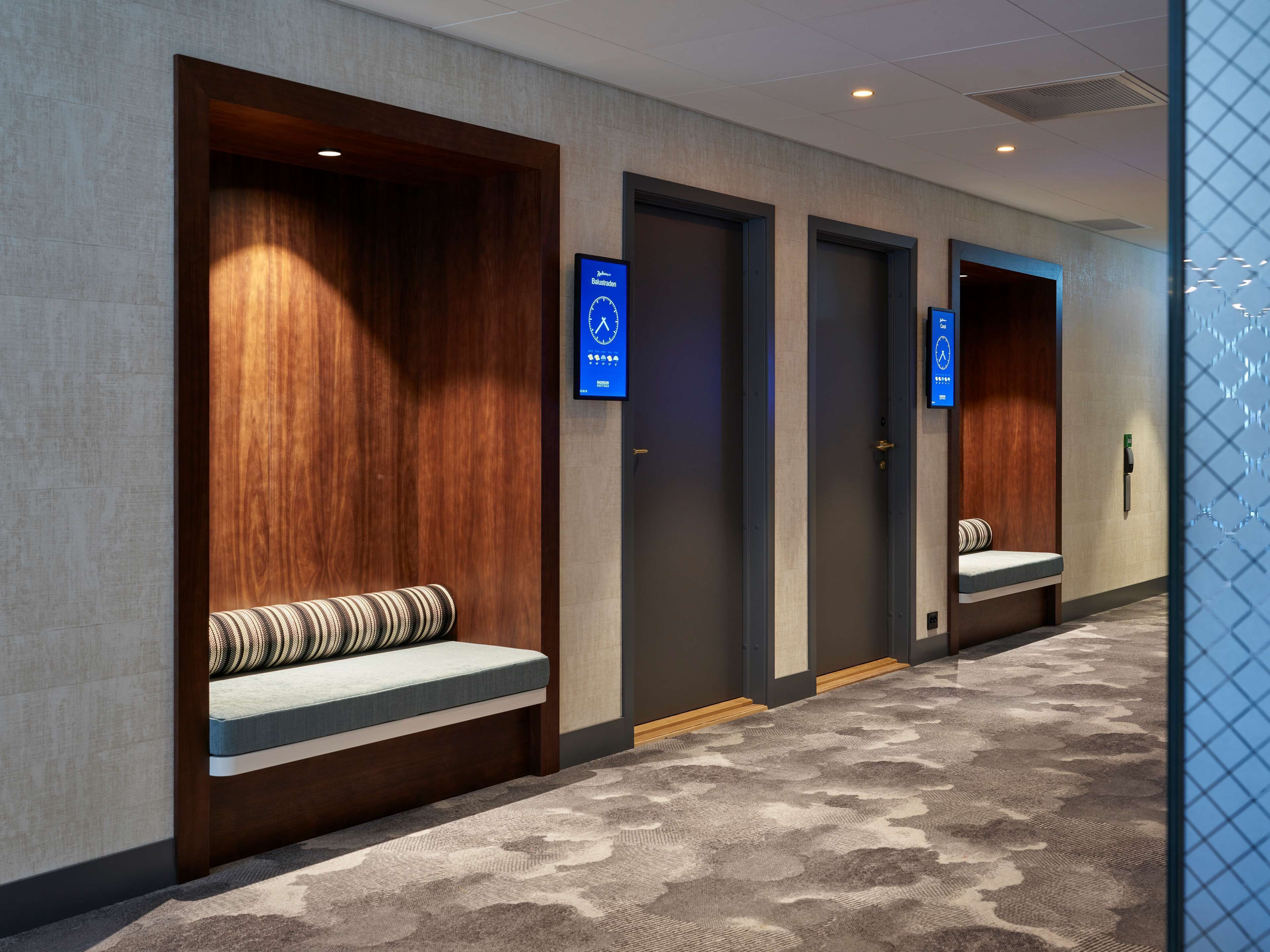 A hotel hallway with wooden paneling, a bench with a striped cushion, and digital clocks on the doors.