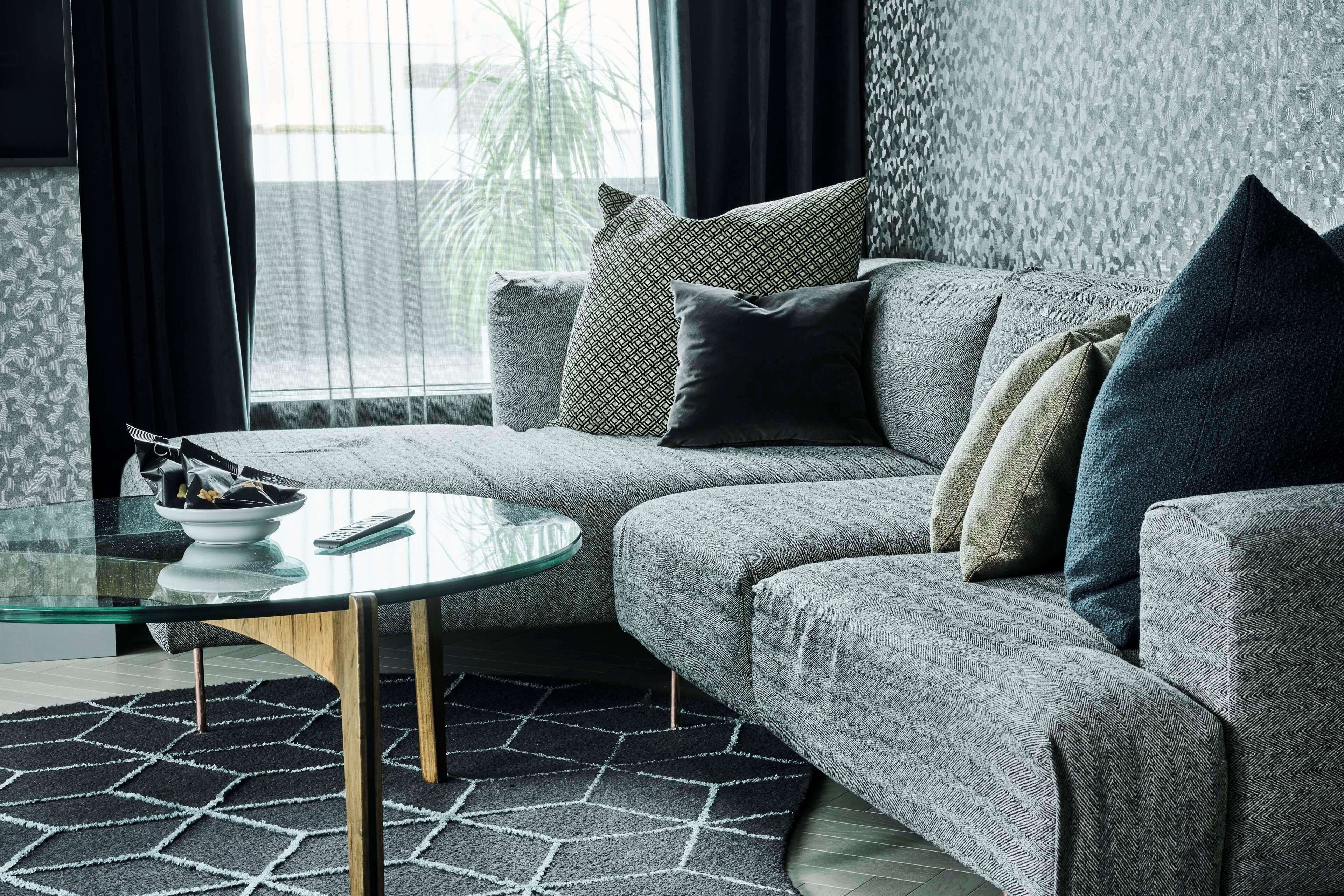 A modern living room with a grey sofa, multiple cushions, a glass coffee table, and a patterned rug.