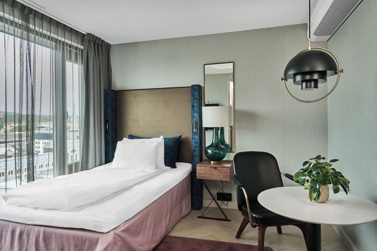 A hotel room with a bed, a desk, a chair, a round table with a plant, and a large window with curtains.