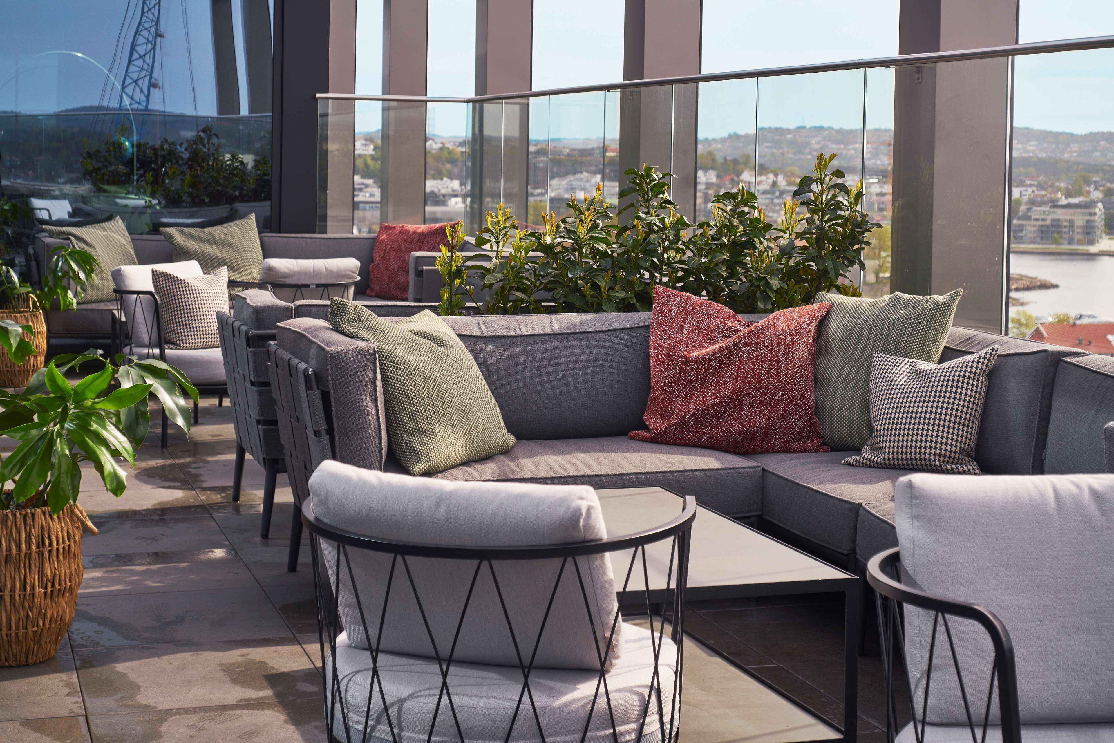Un espacio al aire libre moderno con sofás y sillones, rodeado de plantas en macetas y vistas a un paisaje urbano.