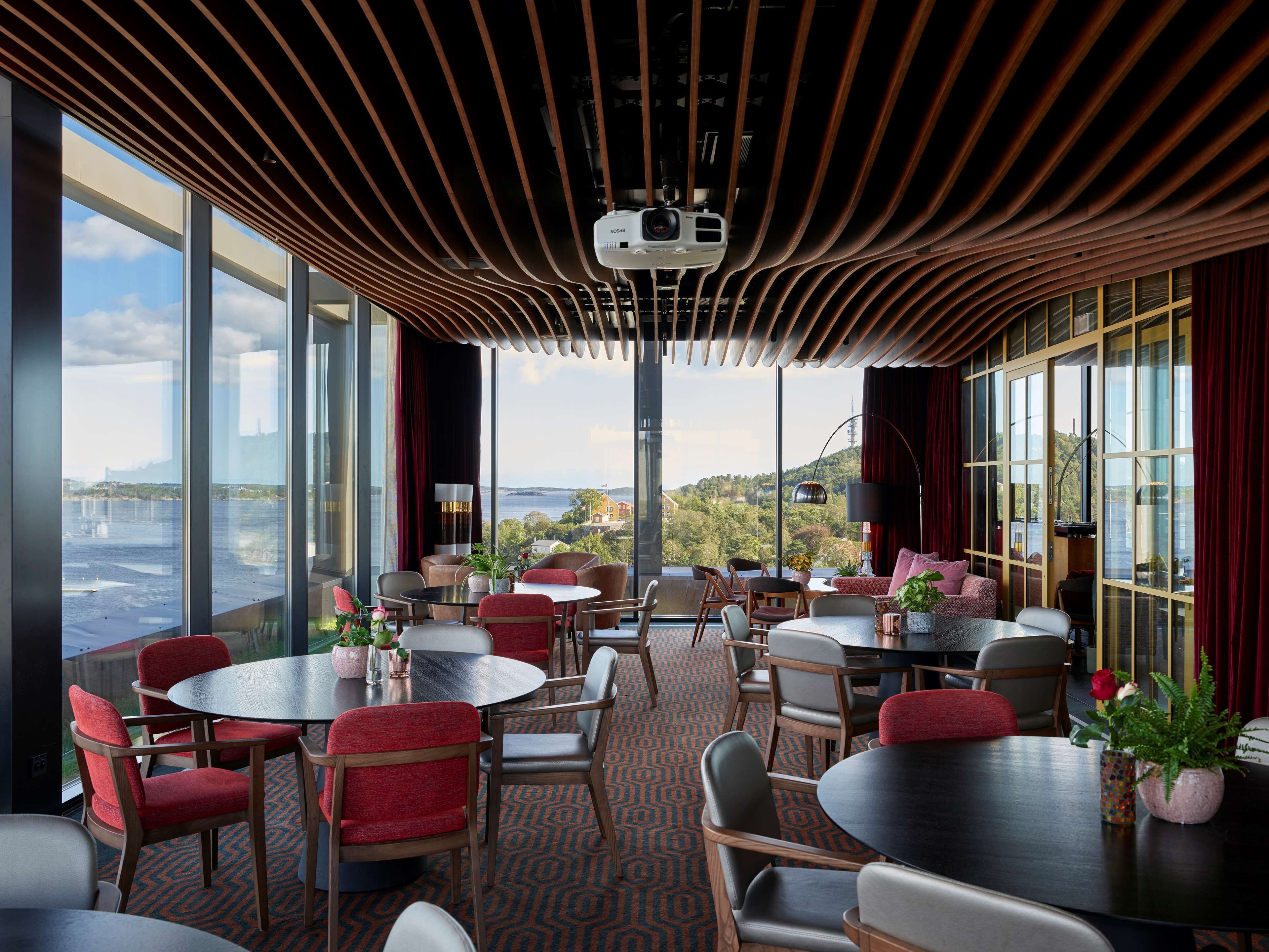 Une chambre d’hôtel moderne dotée de grandes fenêtres offrant une vue sur la ville, d’un coin salon confortable avec des fauteuils rouges et d’un téléviseur à écran plat fixé au plafond.
