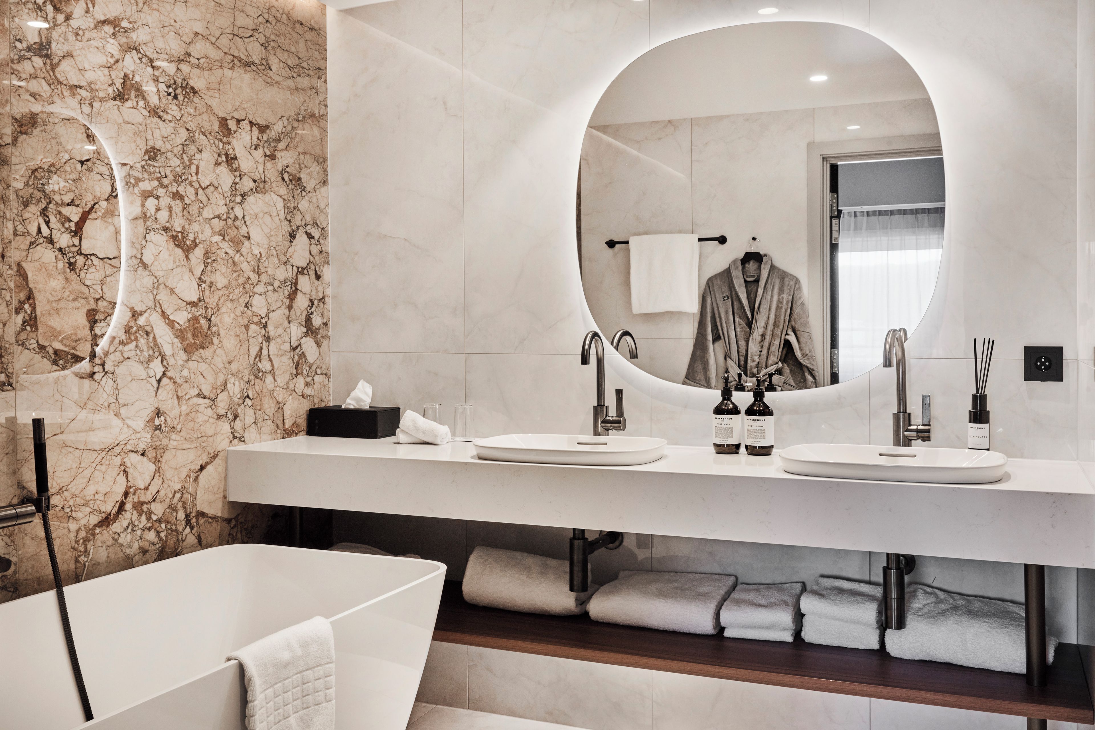 Une salle de bain moderne avec un mur en marbre, deux lavabos, un grand miroir et une baignoire.