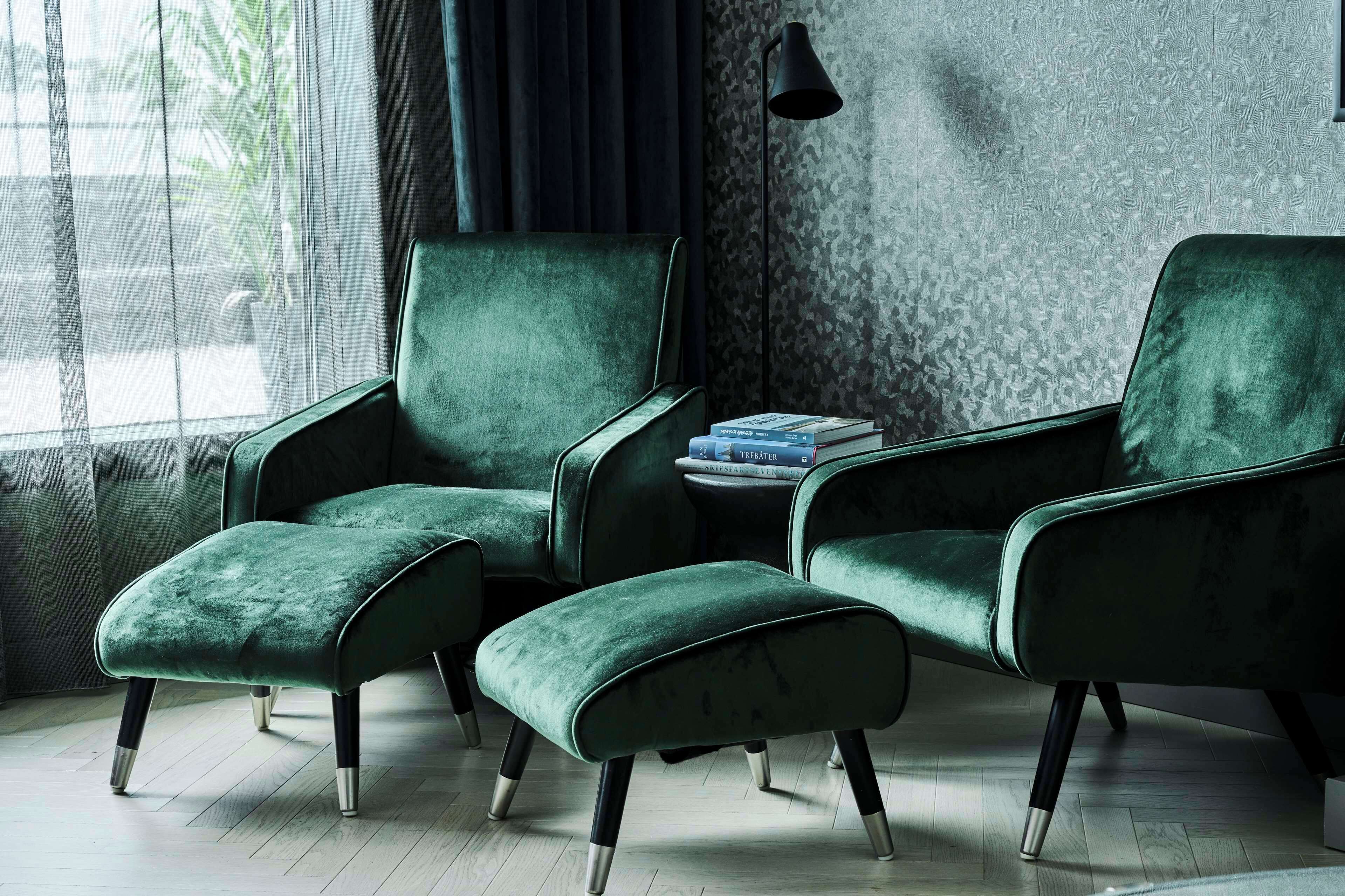 Ein Zimmer mit einem grünen, gepolsterten Sessel und Hocker, einem kleinen Beistelltisch mit Büchern und einer Wand mit Muster.