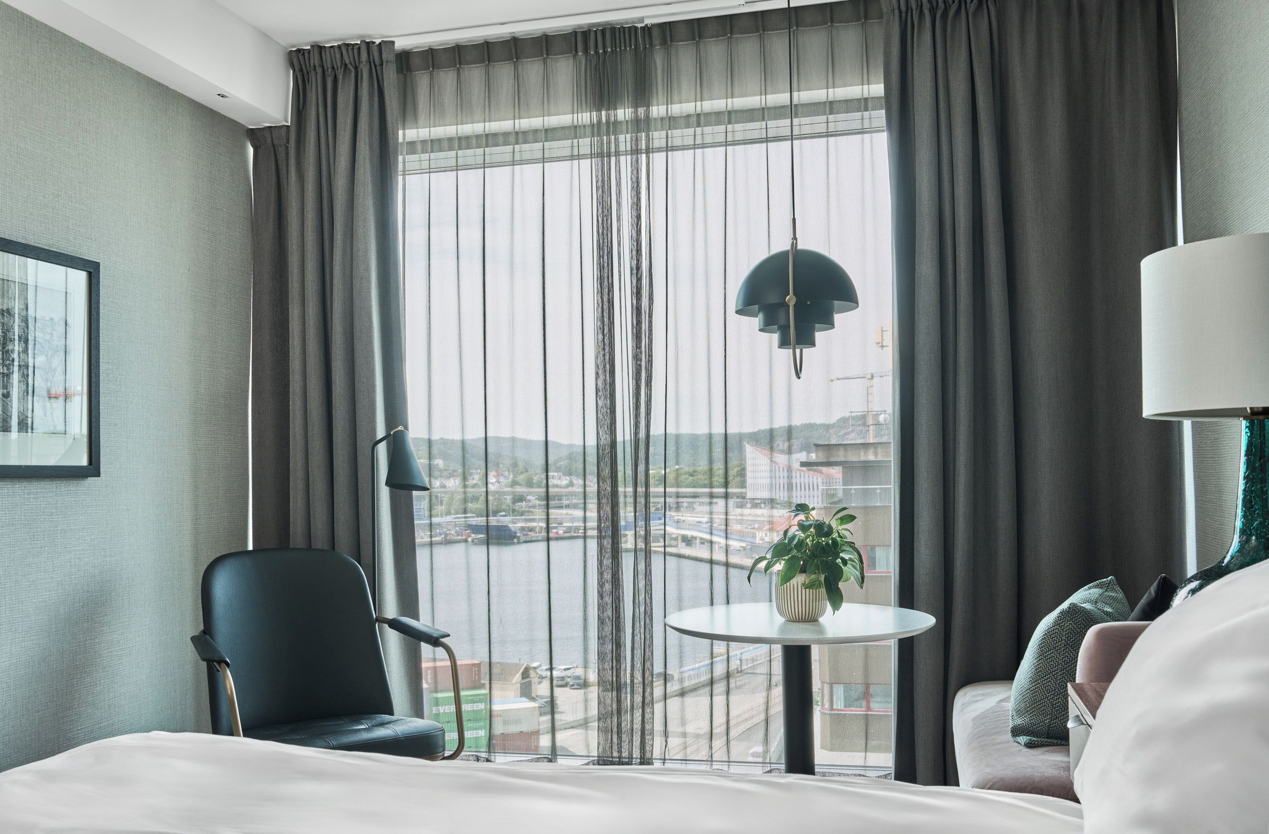 Una habitación de hotel con cama, una silla, una pequeña mesa con una planta, una lámpara y vistas a un paisaje urbano a través de amplios ventanales.