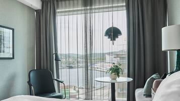 Ett hotellrum med en säng, en stol, ett litet bord med en växt, en lampa och utsikt över en stadspanorama genom stora fönster.