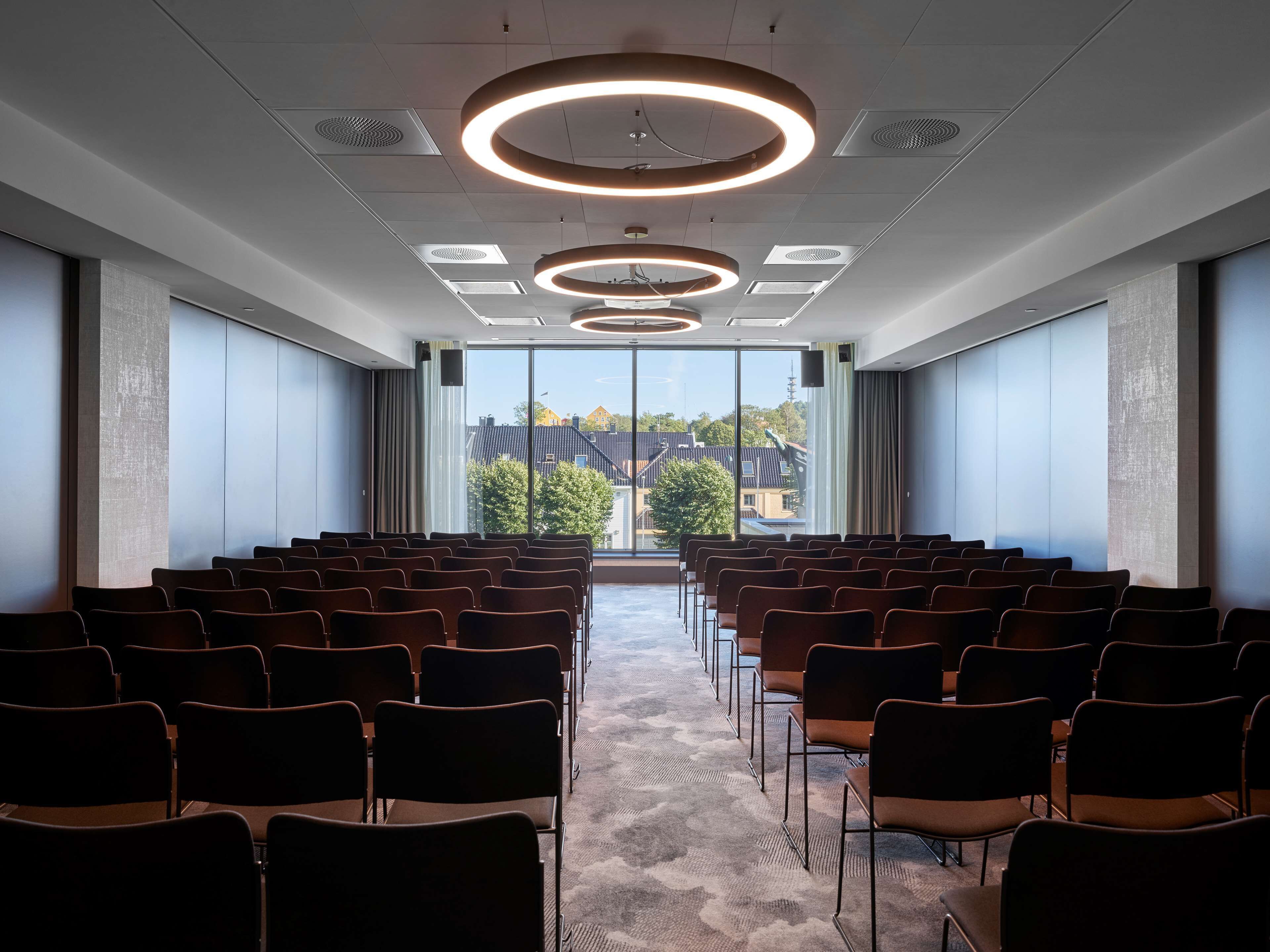 A conference room with rows of chairs, large windows, and circular ceiling lights.