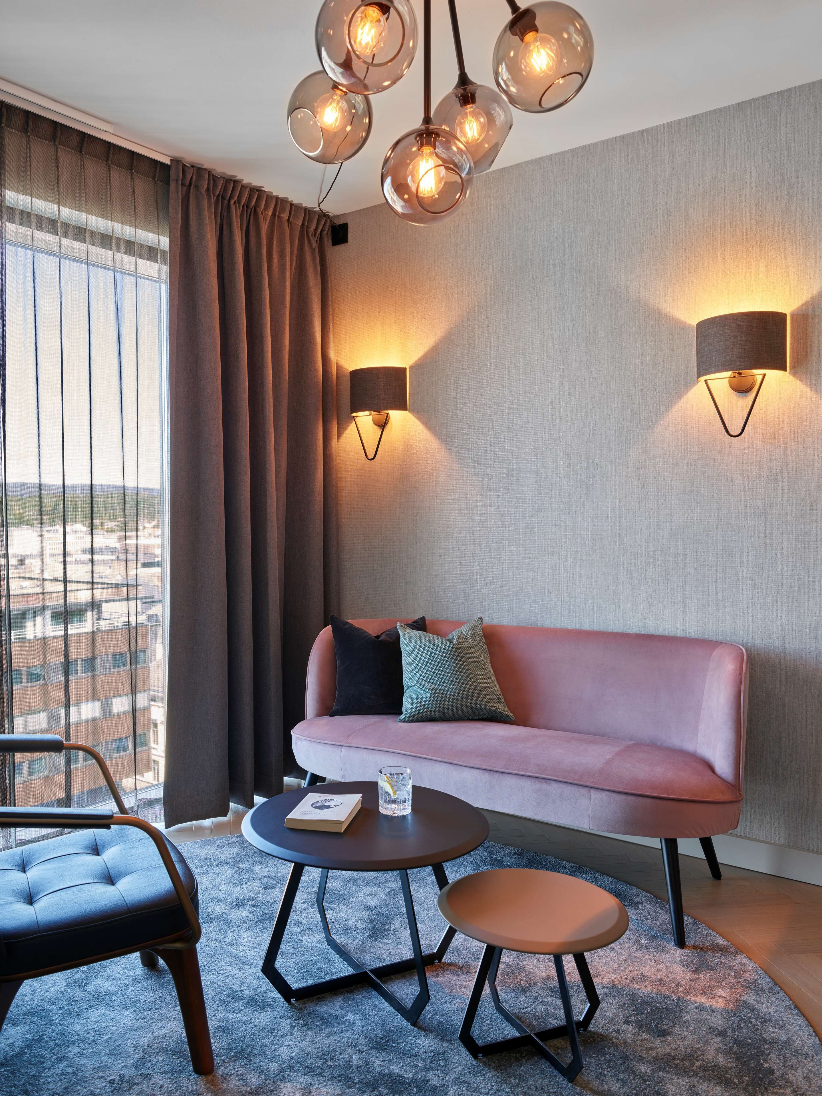 A modern hotel room with a pink sofa, two wall-mounted lamps, a round coffee table, and a view of the city through large windows.