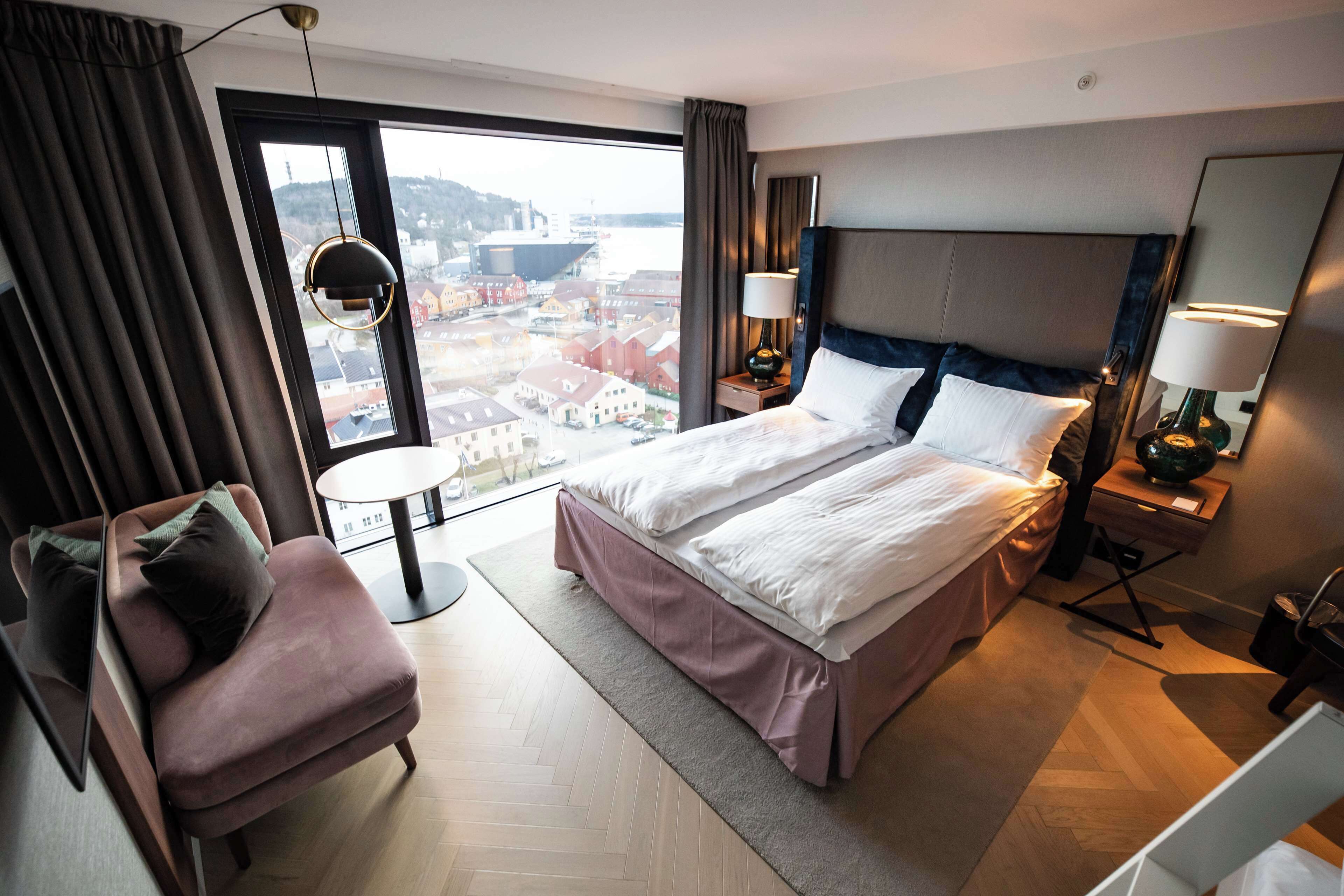 Ein Hotelzimmer mit einem großen Bett, einem Sessel, einem Nachttischchen mit Lampen und Blick auf eine Stadtlandschaft durch ein großes Fenster.
