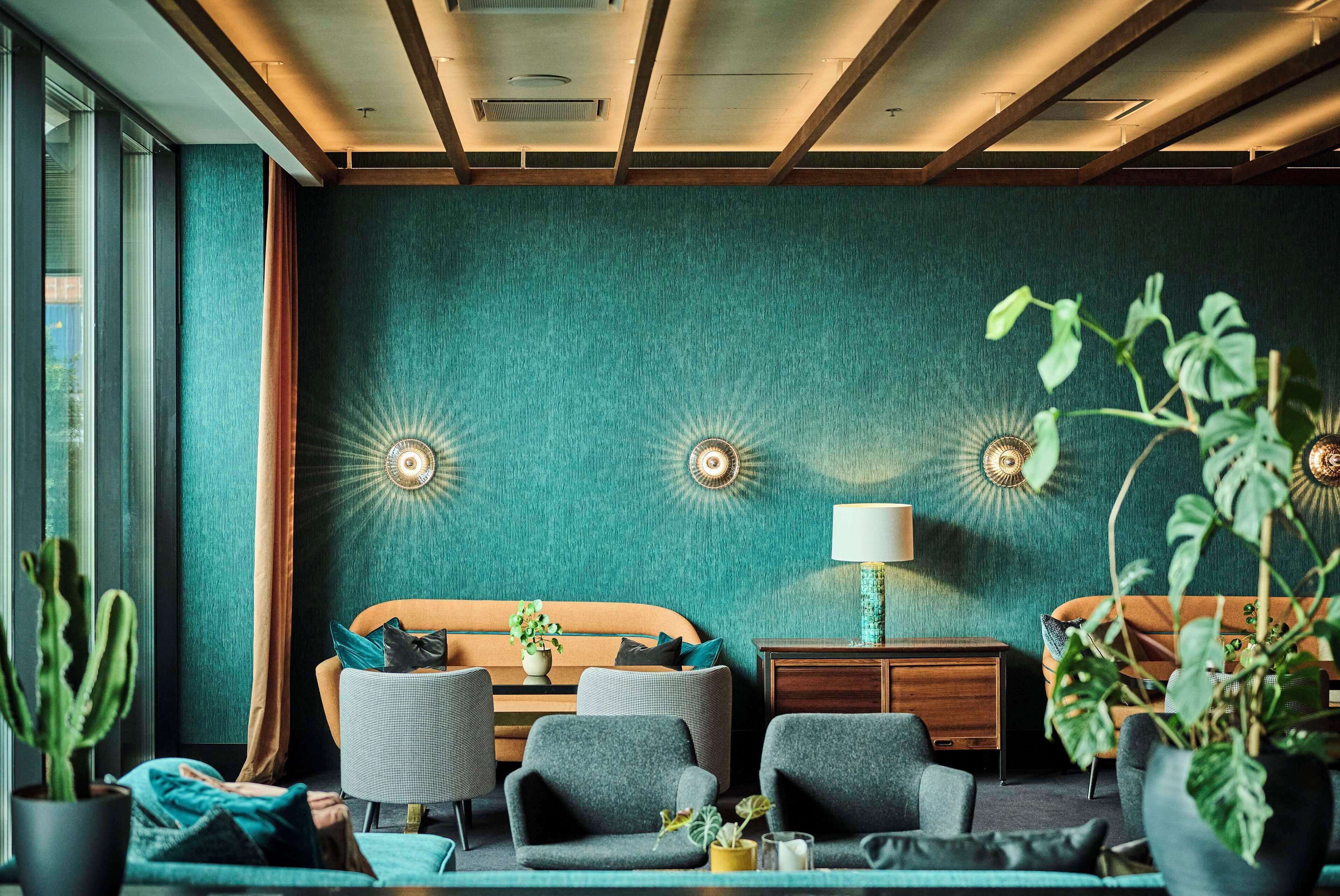 A room with teal walls, a wooden ceiling, a bed with a striped headboard, two armchairs, a small table with a lamp, and a potted plant.