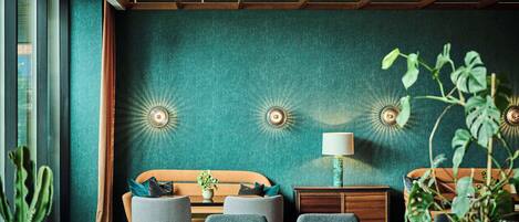 A room with teal walls, a wooden ceiling, a bed with a striped headboard, two armchairs, a small table with a lamp, and a potted plant.