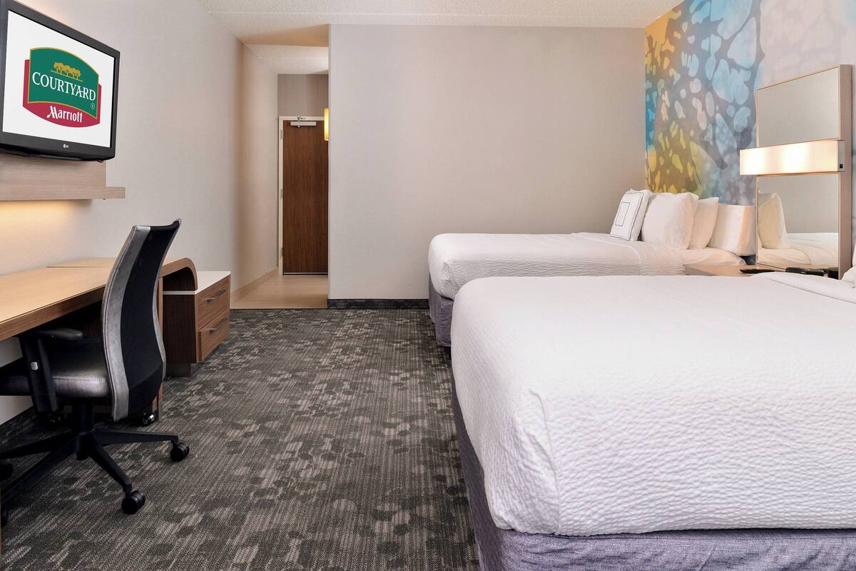 A hotel room with a desk, two beds, a TV displaying the Courtyard Marriott logo, and a painting on the wall.