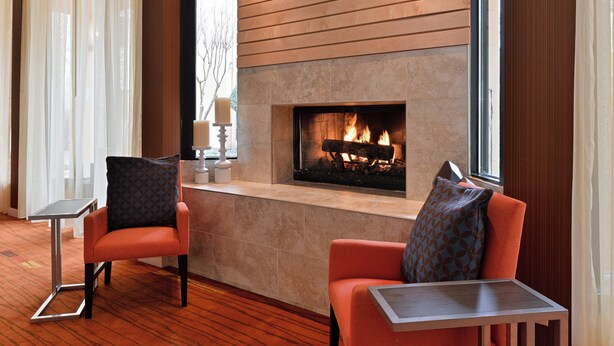 A modern lounge area with a fireplace, two orange chairs, a small table, and a view of the outdoors. - Courtyard by Marriott Dallas Northwest (Dallas)