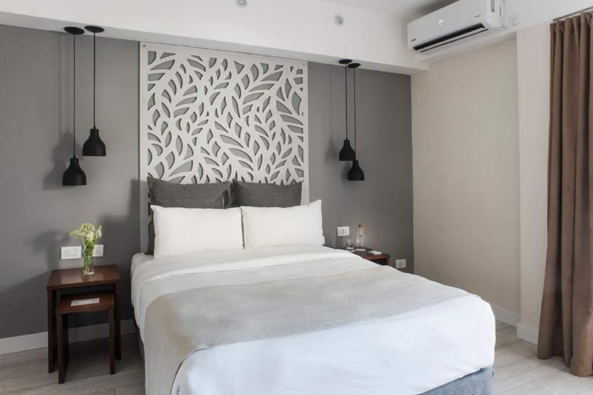 Une chambre d’hôtel moderne avec un grand lit, une tête de lit décorative, deux lampes suspendues, une table de chevet ornée d’un vase de fleurs et un appareil de climatisation.