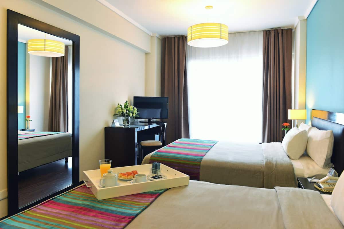 Une chambre d’hôtel avec deux lits, un bureau avec une télévision, un plateau avec le petit-déjeuner et une grande fenêtre avec des rideaux.