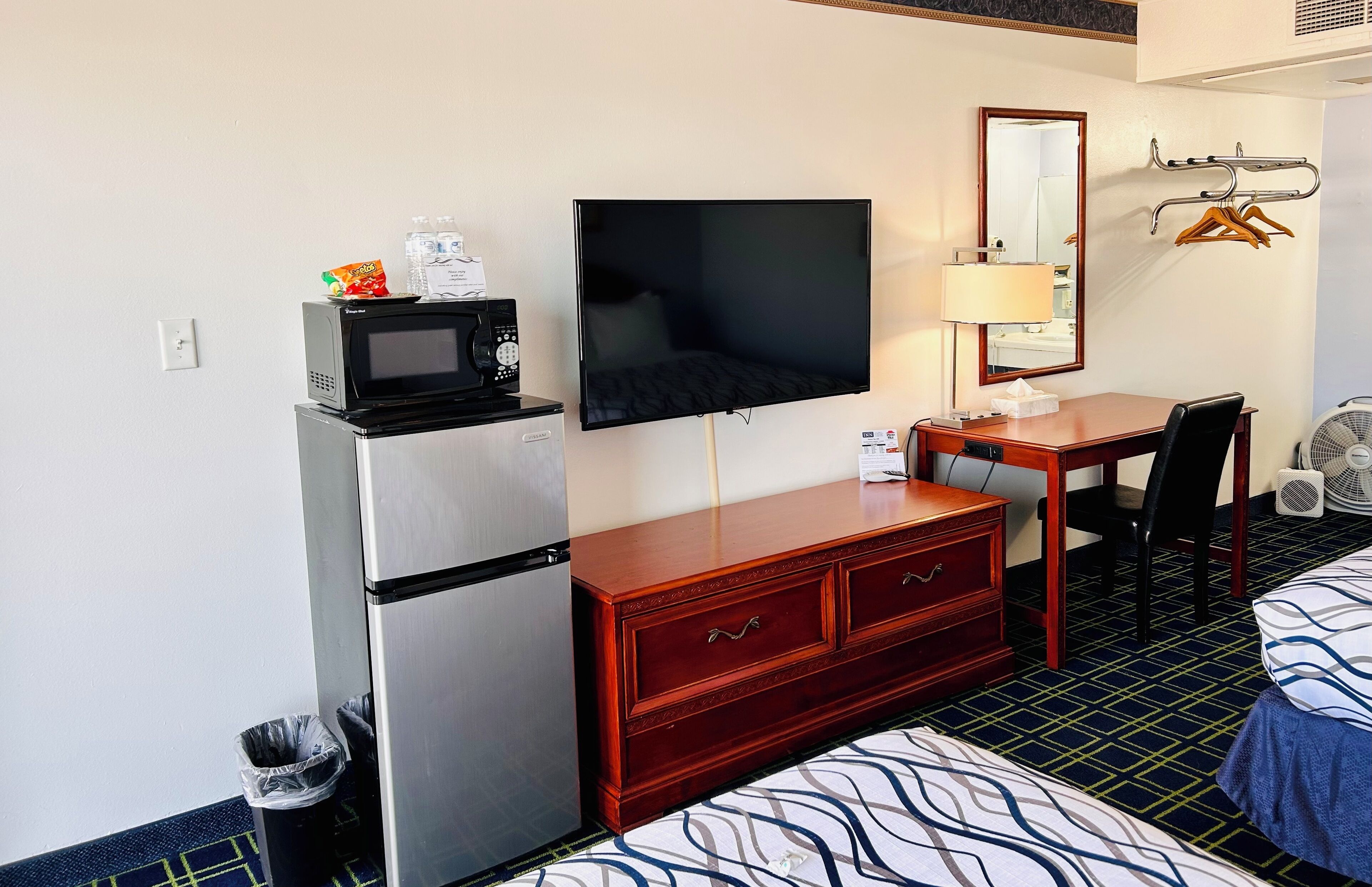 A hotel room with a bed, a desk with a chair, a television, a microwave, a refrigerator, a dresser, a mirror, and a wall-mounted fan.