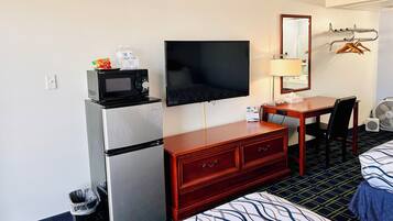 A hotel room with a bed, a desk with a chair, a television, a microwave, a refrigerator, a dresser, a mirror, and a wall-mounted fan.