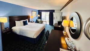 A hotel room with a large bed, a desk, a chair, a desk lamp, a nightstand, a mirror, and a window with curtains. - Hospitality Inn (North Platte)