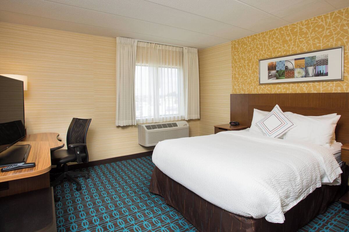 Une chambre d’hôtel avec un lit, un bureau équipé d’un ordinateur, un climatiseur et une fenêtre avec des rideaux.