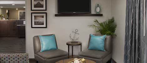 A lobby with two grey sofas, a round wooden coffee table, a small side table, a flat-screen TV, framed pictures, and a potted plant.