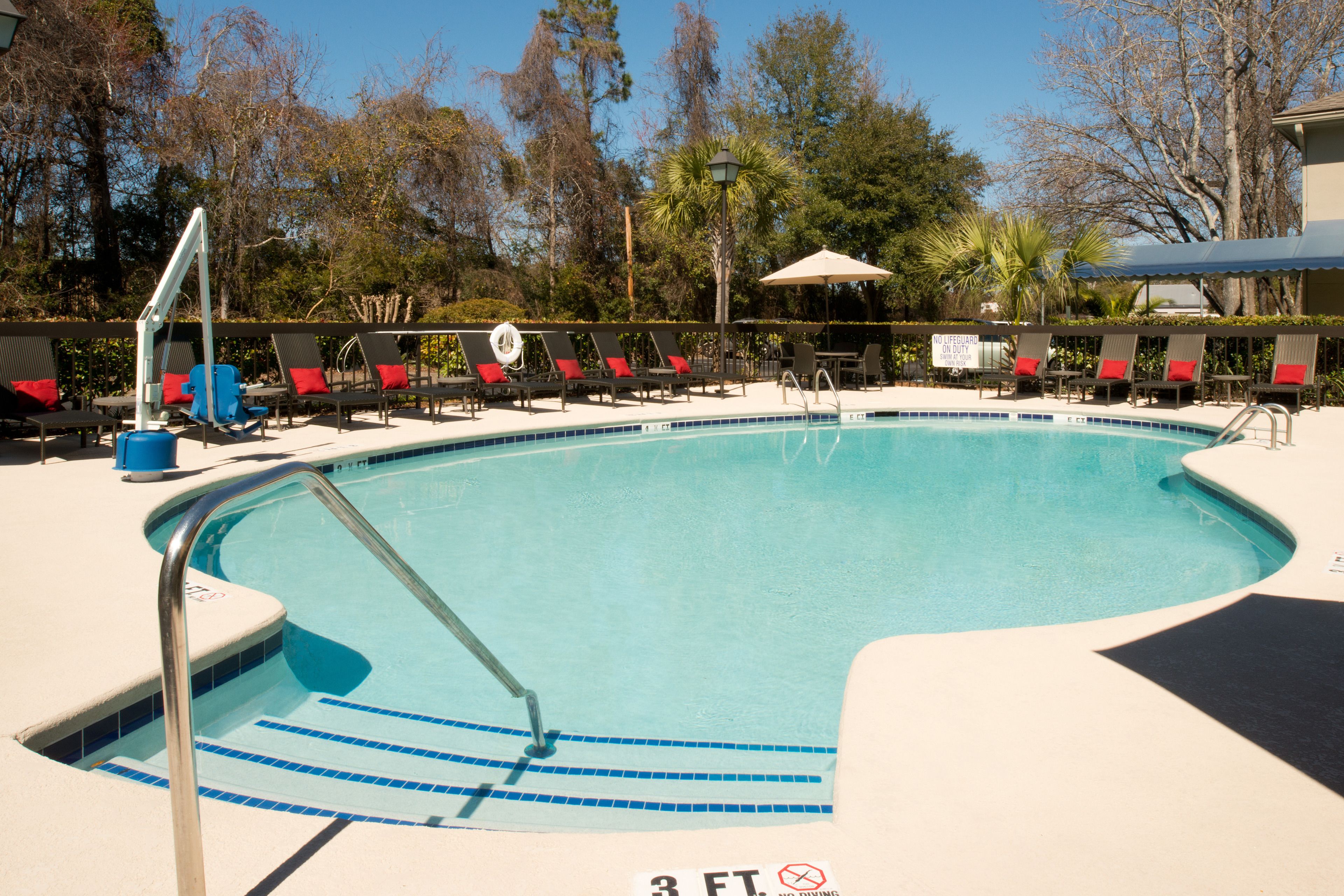 A swimming pool with a 3 ft depth marker, surrounded by lounge chairs with red cushions, a palm tree, and a covered seating area.