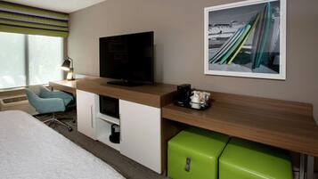 Quarto de hotel com uma cama, uma escrivaninha com televisão de tela plana, um minibar verde, uma cadeira e um quadro na parede.