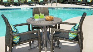 Zona junto a la piscina con sillones de mimbre y una mesa, piscina y sillones de descanso al fondo.