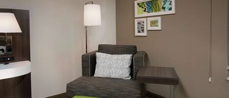 A hotel room with a grey armchair, a small table, a green ottoman, a lamp, and artwork on the wall.
