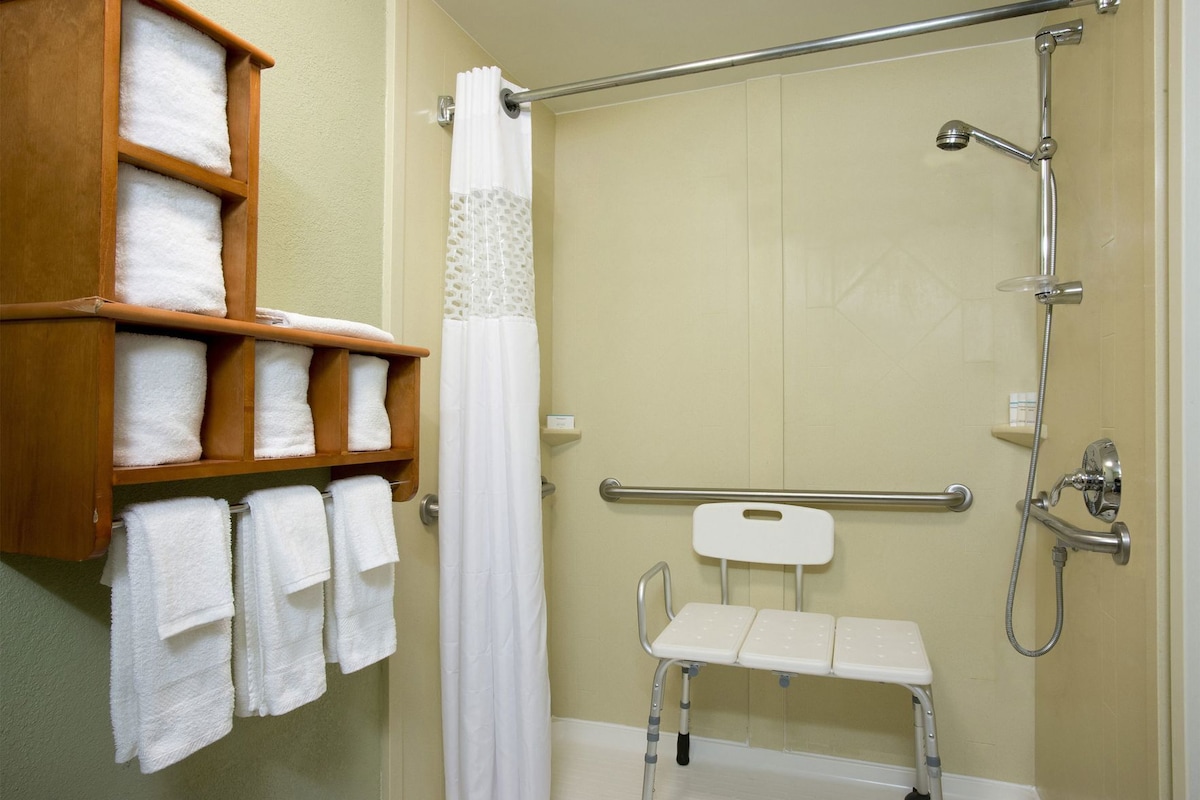 A shower area with a grab bar, a white shower curtain, and a shelf with towels.