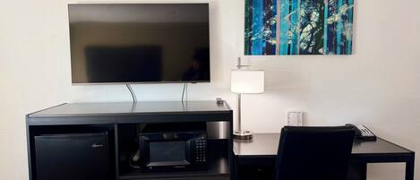 A compact hotel room with a flat-screen TV, a microwave, a desk with a chair, a lamp, and a trash bin.
