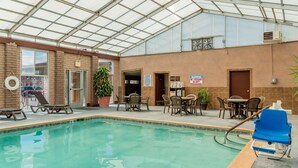 An indoor swimming pool with lounge chairs and a table, surrounded by brick walls and a glass ceiling.