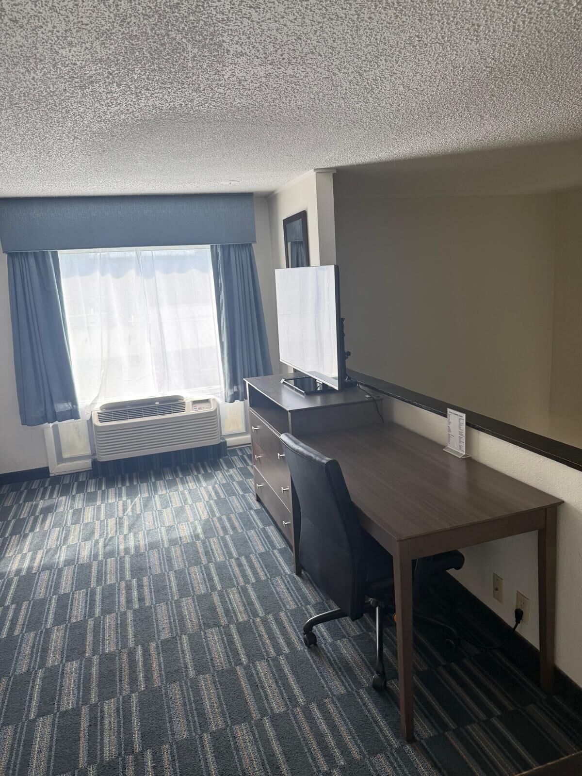 A hotel room with a desk, chair, air conditioning unit, and a large window with curtains.