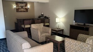 A hotel room with a sofa, armchairs, a small table, a TV, and a desk with a lamp.