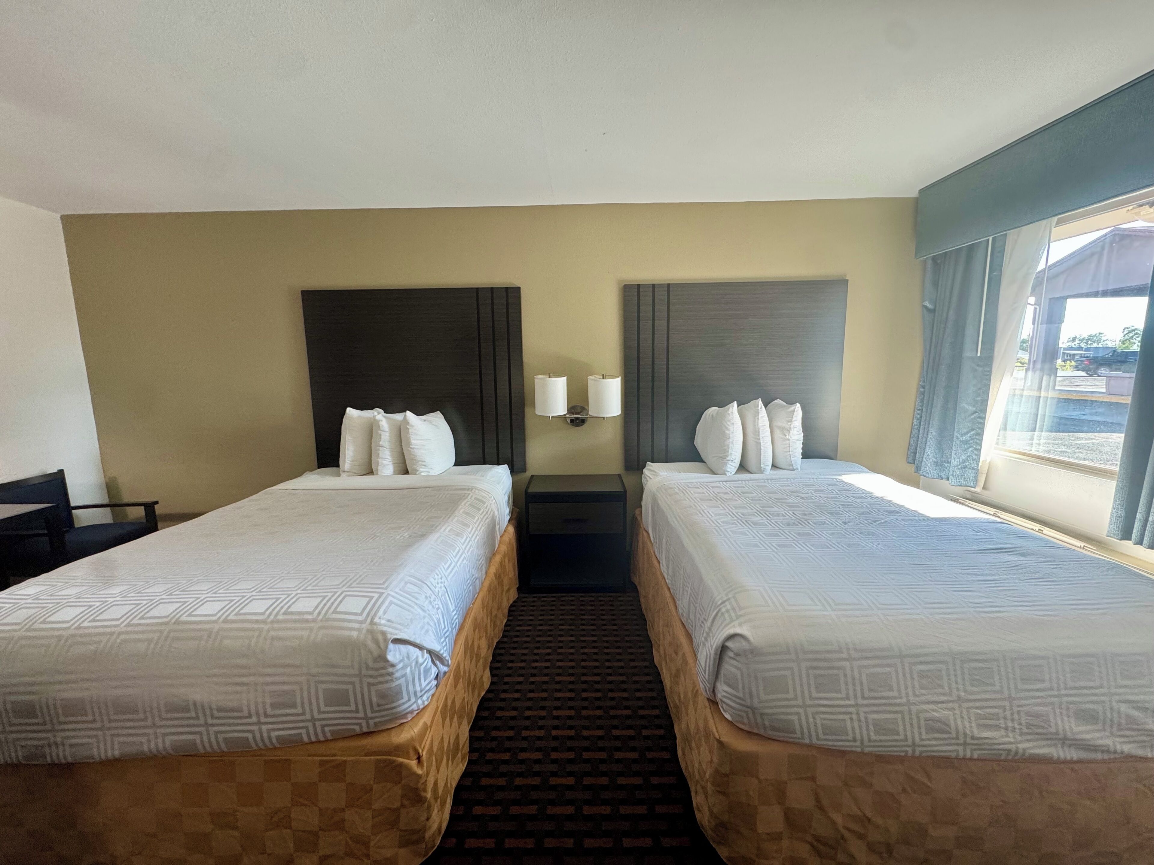 A hotel room with two beds, a window with curtains, a wall-mounted lamp, and a checkered carpet.