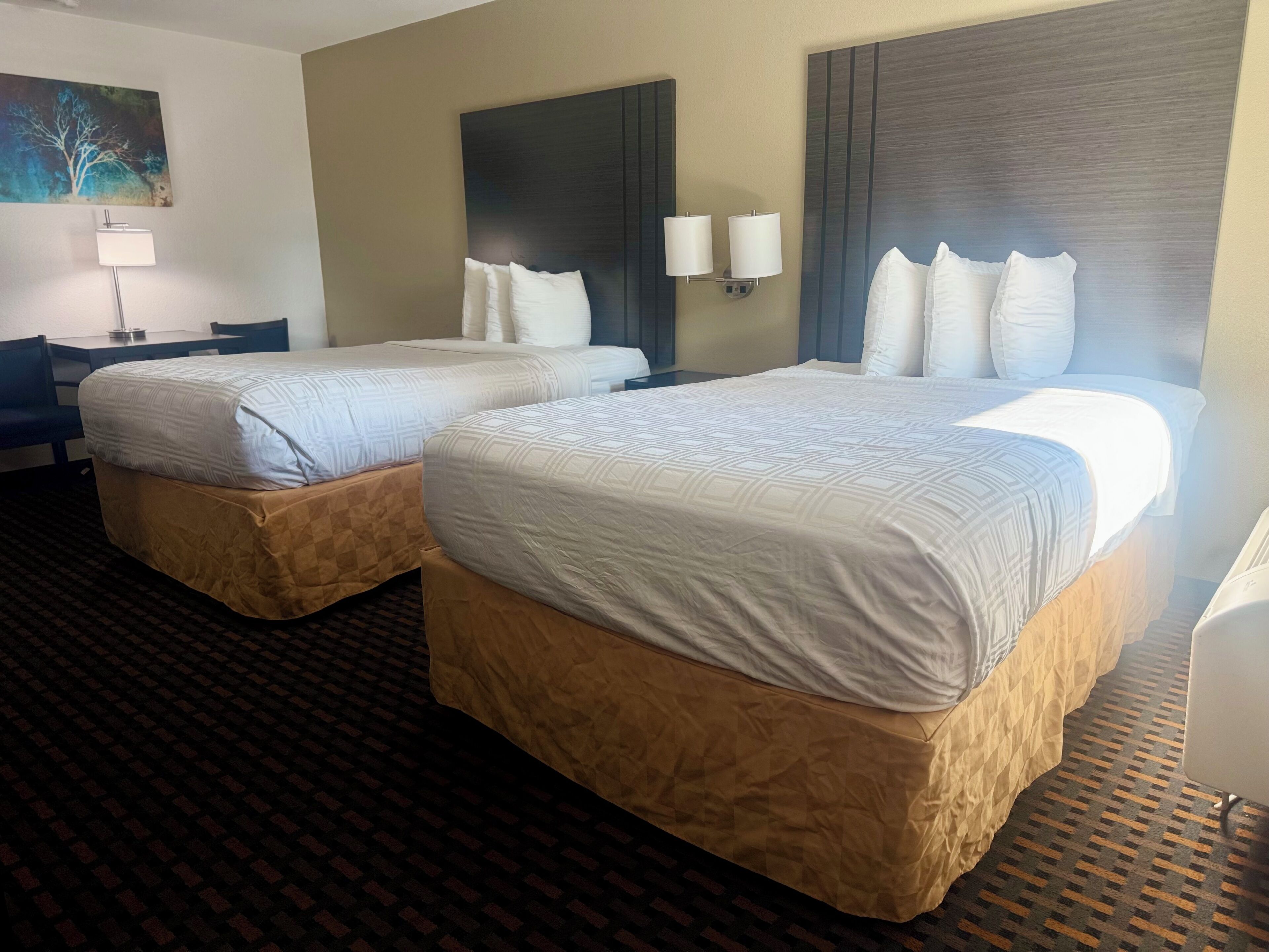A hotel room with two beds, a desk, a chair, a lamp, and a painting on the wall.
